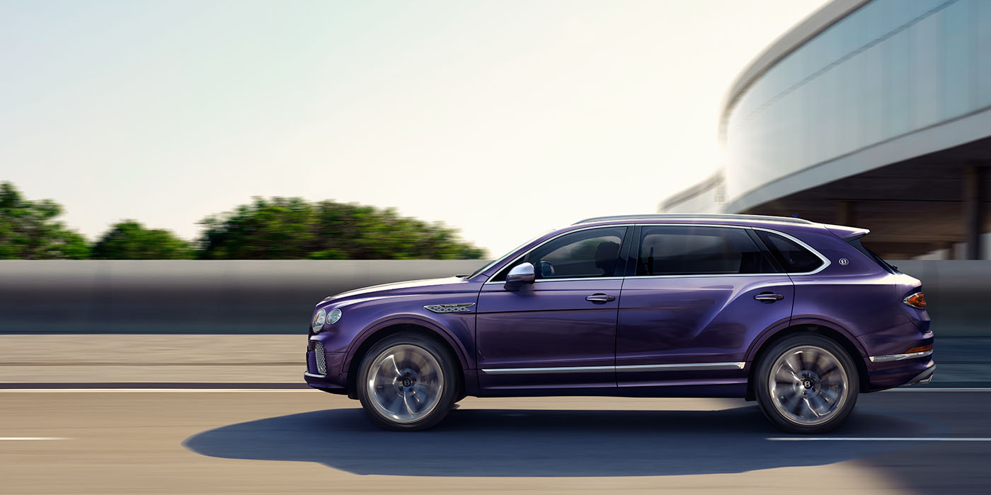 Bentley Muscat Bentley Bentayga Extended Wheelbase Mulliner luxury SUV profile in Tanzanite Purple paint, driving on an country road past trees.