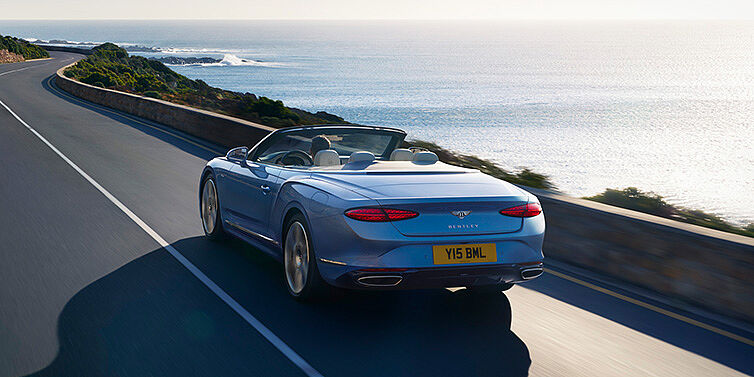 Bentley Muscat New Bentley Continental GTC Azure convertible rear view in Light Windsor blue paint with 22 inch ten spoke wheels driving dynamically on an ocean road in the sunshine