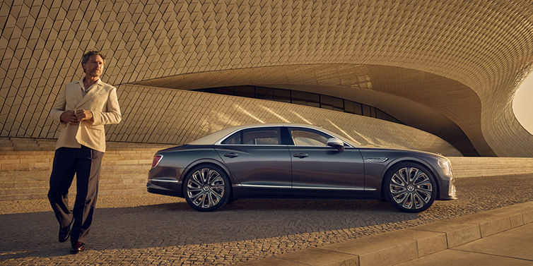 Bentley Muscat Bentley Flying Spur Mulliner sedan in Rose Gold over Magnetic duotone paint parked by a curved wall with person in suit jacket standing on the left